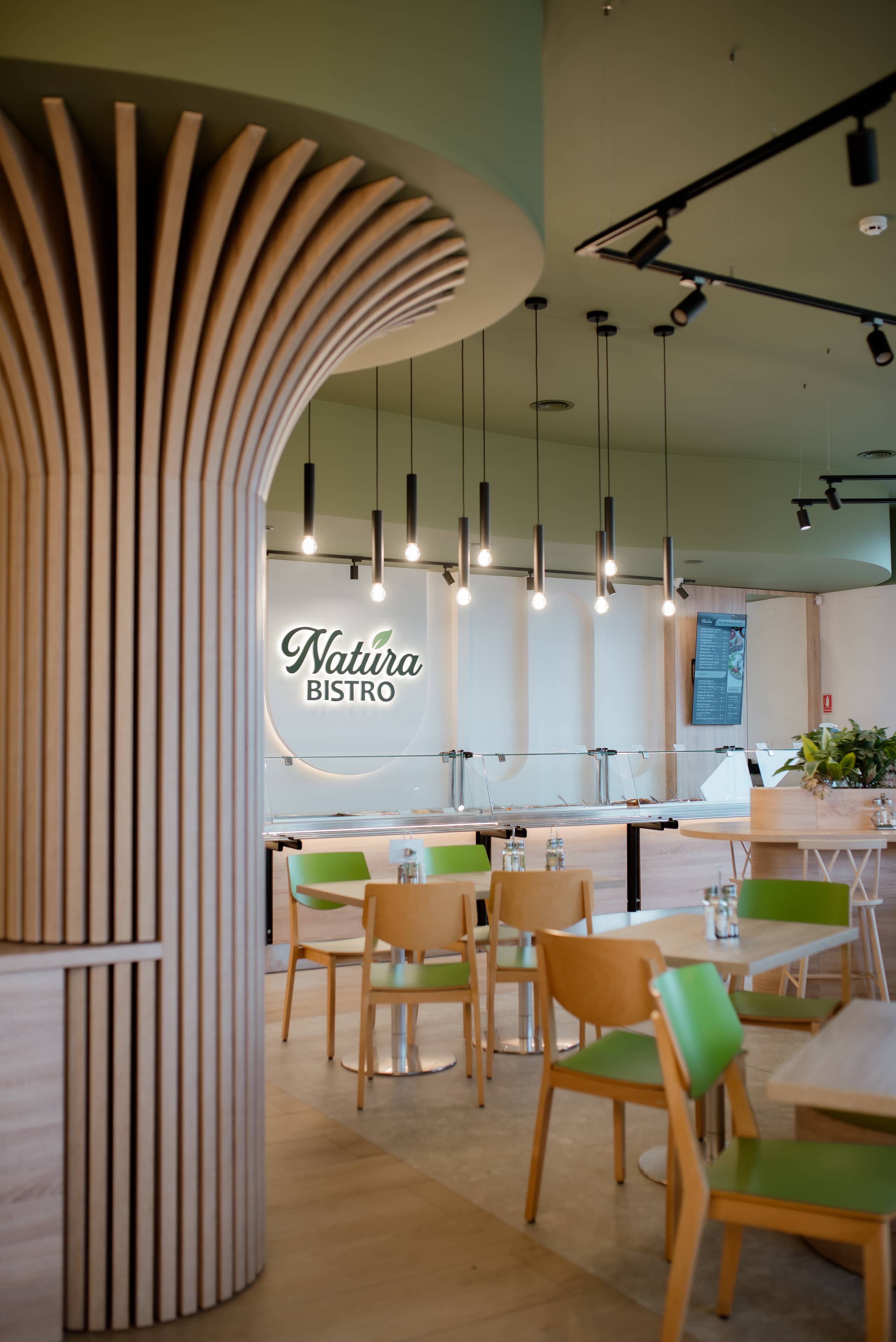 Sala unui bistro cu coloană canelată din lemn deschis, mese de lemn și tapițerii verzi